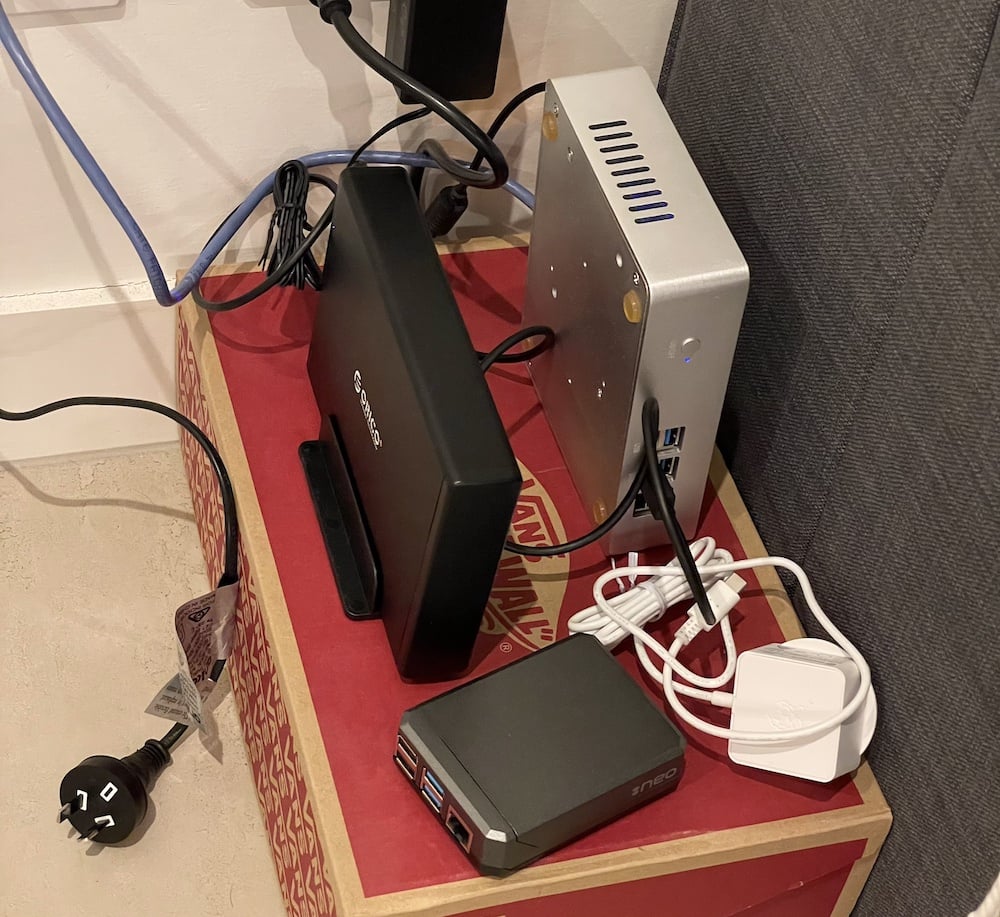 a small computer sitting on a shoebox with an external HDD next to it, surrounded by a nest of cables